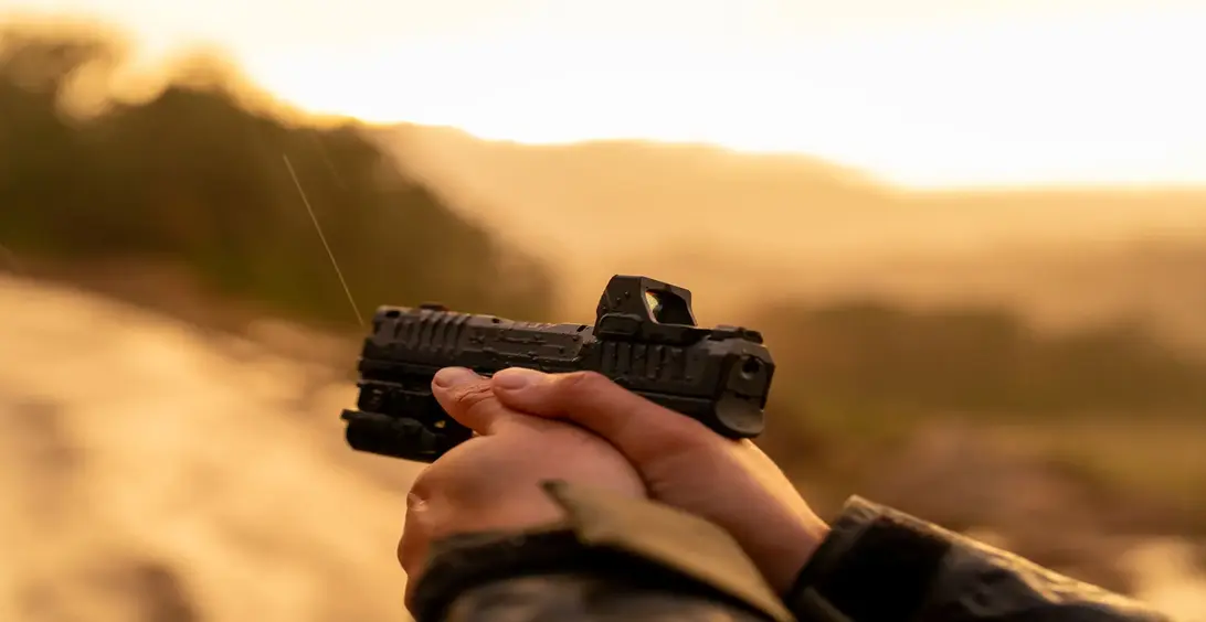 Person aiming a handgun with a sight at sunset.